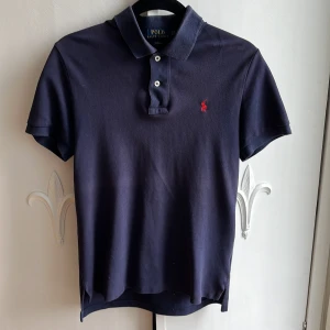 Marinblå pikétröja Polo Ralph Lauren - Klassisk marinblå pikétröja från Polo Ralph Lauren i slim fit-modell. Tröjan har korta ärmar, krage och knäppning med två knappar samt den ikoniska röda logotypen broderad på bröstet. Tillverkad i mjuk bomull som är skön mot huden den har inga defekter och passar dig om du e runt 160/170 cm 