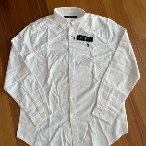 Vit, långärmad Ralph Lauren-skjorta - *   Ralph Lauren-skjorta *   Storlek: L *   Vit med marinblå logga *   Helt ny – aldrig använd  Stilren och oanvänd skjorta från Ralph Lauren i storlek L. Perfekt för en avslappnad och sofistikerad stil. Den klassiska marinblå loggan är broderad på bröstet. Levereras med alla originaletiketter. Ett utmärkt tillskott till din garderob för en tidlös look!  Pris kan diskuteras. Kontakta mig om ni vill ha fler bilder