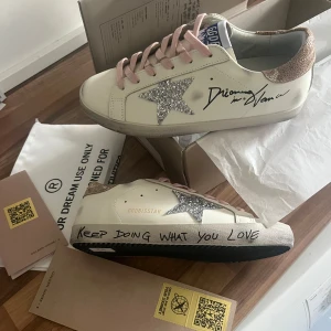 GGDB sneakers med glitterstjärna, strl 40 - Snygga vita sneakers från Golden Goose med rosa snören, silverglittrig stjärna på sidan och beige hälparti med struktur. Textdetaljer som 'Keep doing what you love' på sulan och handskriven stil på sidan. Perfekta för dig som gillar unika detaljer och cool design. Kvitto finns.