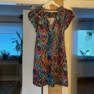 Färgglad kortklänning från Mantaray - Säljer en superfin kortklänning från Mantaray med färgstarkt bladmönster i blått, orange, rosa och svart. Klänningen har v-ringning, korta ärmar och knyt i midjan. Perfekt för dig som gillar unika prints och vill sticka ut lite extra.