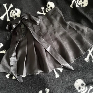 Svart handsydd minikjol med rosett - Supersnygg svart plisserad kortkjol med dekorativa knappar och en stor satinrosett framtill. Kjolen har dragkedja i sidan och detaljer i spets. Perfekt för dig som gillar alternativ stil. Plagget är handsytt, vilket innebär att det kan förekomma mindre skavanker tex lösa trådar! 🪡 