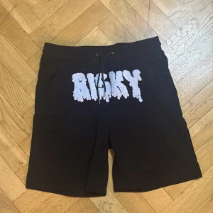 Svarta shorts från Risky med brodyr - Svarta shorts från Risky med stor vit broderad text framtill. De har snörning i midjan och en loose fit som ger en avslappnad vibe. Perfekta för sommaren och streetwear-stilen. Materialet känns mjukt och bekvämt, och designen sticker verkligen ut.