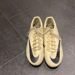 Nike mecurials fotbollsskor i beige och svart - Pris kan diskuteras Snygga fotbollsskor från Nike i beige med svarta detaljer och stor Swoosh-logga. Skorna har snörning, platt sula med dobbar och är tillverkade i syntetmaterial. Perfekta för dig som vill sticka ut på planen med stilren design.