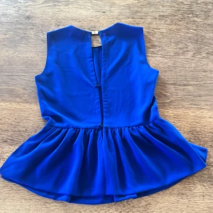 Zara Kornblå ärmlös blus med volang - Supersnygg kornblå blus i glansigt tyg med ärmlös design och rund halsringning. Toppen har en volangliknande peplum nedtill och en öppen detalj i ryggen med knäppning. Perfekt för dig som vill sticka ut med färg och stil. Tryck ej köp nu