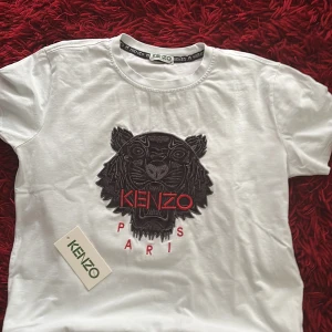 Kenzo t-shirt - Snygg vit t-shirt från Kenzo med ikonisk svart tigerbrodyr och röd text på bröstet. Klassisk passform och rund halsringning. Perfekt statement-plagg för dig som vill sticka ut med stilren design och coolt tryck.