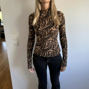 Zebramönstrad polotröja i brunt och svart - Säljer en tight zebramönstrad polotröja i brunt och svart. Tröjan har lång ärm, hög krage och sitter slimmat mot kroppen. Perfekt för dig som gillar att sticka ut med coola mönster och vill ha en trendig look.