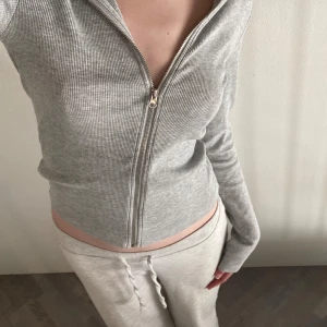 Grå ribbad zip hoodie - Säljer en ljusgrå ribbad hoodie med dragkedja framtill. Tröjan är croppad och har långa ärmar. Perfekt för en chill och avslappnad stil. Materialet är mjukt och stretchigt, troligtvis bomull eller bomullsblandning.