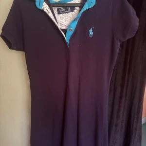 Mörkblå kortklänning Polo Ralph Lauren - Snygg mörkblå kortklänning från Polo Ralph Lauren med turkosa detaljer vid kragen och knappar framtill. Klänningen har korta ärmar och en klassisk pikékrage, samt det ikoniska broderade logotypen på bröstet.