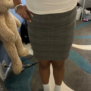 Grå rutig pennkjol - Snygg grå pennkjol med klassiskt rutigt mönster. Kjolen är kort och har en tight passform som framhäver figuren. Perfekt för dig som gillar en stilren och trendig look.