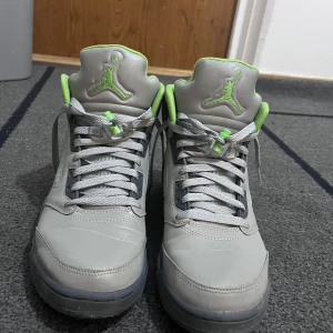 Nike Air Jordan 5 Retro GREEN BEAN - Säljer ett par Nike Air Jordan 5 Retro i grått med limegröna detaljer. Skorna har hög siluett, snörning och coola meshpaneler på sidorna. Insidan och loggan är limegröna, sulan är grå med limegrön detalj. Perfekta för dig som vill sticka ut med din sneakerstil.