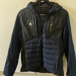 Mörkblå Sail Racing dunjacka S - Säljer en mörkblå dunjacka från Sail Racing med broderade loggor och patchar från Antarctica Expedition. Jackan har huva, dragkedja framtill och flera praktiska fickor. Perfekt för kalla dagar och snygg streetstil.