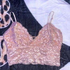 Rosa spetsig bralette topp - Superfin ljusrosa bralette topp med blommigt spetsmönster och tunna justerbara axelband. Toppen har en omlottliknande v-ringning och är fodrad på insidan för extra komfort. Perfekt för att styla under en skjorta eller som den är till jeans.