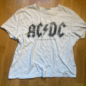 Vit AC/DC t-shirt - Vit t-shirt med stort AC/DC-tryck och texten 'Let There Be Rock 1977' på bröstet. Klassisk rund hals och korta ärmar. Skön passform i mjuk bomull, perfekt för dig som gillar bandtröjor och rockstil. 💕