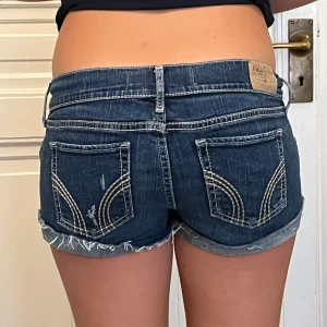Mörkblå jeansshorts från Hollister - Snygga mörkblå jeansshorts från Hollister med låg midja, slitna detaljer och råa kanter. Shortsen har dubbla knappar framtill, klassiska bakfickor med vit broderad söm och uppvikta benslut. Perfekta för varma dagar och en avslappnad look.