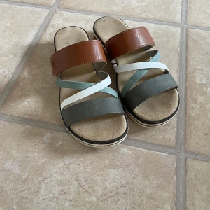 Färgglada sandaler från Rieker - Snygga sandaler från Rieker med breda remmar i brunt, grått, vitt och ljusgrönt. Skorna har platt sula och öppen tå, samt en bekväm fotbädd. Perfekta för varma dagar när du vill ha både stil och komfort. Bra skick då de är sparsamt använda.