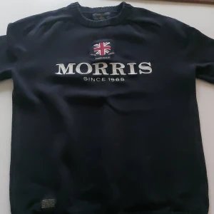 Mörkblå stickad tröja Morris Gents XS - Snygg mörkblå stickad tröja från Morris Gents med broderad Union Jack och tydlig logga framtill. Klassisk rund halsringning och långa ärmar. Perfekt för dig som gillar stilren och tidlös design. 150-165CM