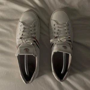 Vita Moncler sneakers med röd/blå rand - Snygga vita sneakers från Moncler i skinn med klassiska röda och blå ränder på sidan. Skorna har rund tå, platt sula och snörning framtill. Moncler-logga på plösen ger en exklusiv touch. Perfekta för dig som gillar stilrena och sportiga skor. Lite fläckar på under sulan på vänstra skon. Inget og medföljer skriv i pm för mer bilder :)  