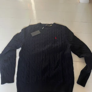 Mörkblå kabelstickad tröja Polo Ralph Lauren - Säljer en mörkblå kabelstickad tröja från Polo Ralph Lauren med klassisk röd broderad logga på bröstet. Tröjan har rund halsringning, långa ärmar och ribbade muddar. Perfekt för dig som gillar stilren och tidlös design.