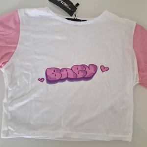 Vit och rosa t-shirt  - Framsidan har ett tryck med ordet BABY i bubbliga rosa bokstäver och små hjärtan. T-shirten är kortärmad och har en croppad passform, perfekt till jeans eller kjol.