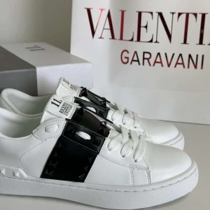 Valentino Garavani Skor - Snygga vita Valentino Rockstud sneakers i skinn med svart detalj och ikoniska nitar över vristen. Klassisk rund tå, platt sula och snörning. Perfekta för dig som vill sticka ut med en lyxig och edgy vibe.