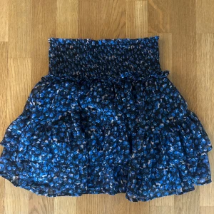 Blommig blå volangkjol från Neo Noir - Supersöt kort volangkjol från Neo Noir med blått och svart blommönster. Kjolen har smock i midjan för en bekväm passform och flera lager volanger. 