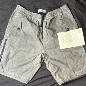 Grå shorts från Stone Island -  Stone Island Reflective shorts. elastisk midja och snörning. De har två framfickor med knappdetaljer och dragkedjor på sidorna. Baksidan pryds av en stor Stone Island-logga. Perfekta för sommaren och riktigt sköna att bära.