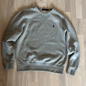 Grå sweatshirt från Polo Ralph Lauren - Snygg grå sweatshirt från Polo Ralph Lauren med klassisk rund hals och broderad logga på bröstet. Tröjan är i mjuk bomullsmix och har ribbade muddar vid ärmslut och midja. Perfekt för en chill och stilren look.