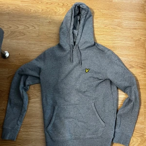 Grå hoodie från Lyle & Scott - Snygg grå hoodie från Lyle & Scott med klassisk gul logga på bröstet. Tröjan har huva med snörning, känguruficka och ribbade muddar. Perfekt för chill dagar eller när du vill ha en enkel och stilren look.