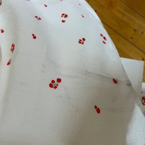 Vit långklänning med blommigt mönster - Superfin långklänning från Adoore i vit med små röda blommor. Klänningen har v-ringning, knappar framtill och långa puffärmar med resår vid handlederna. Materialet känns lätt och flowy. Har några svarta märken ibak men som inte är jätte synliga. Endast använd en gång