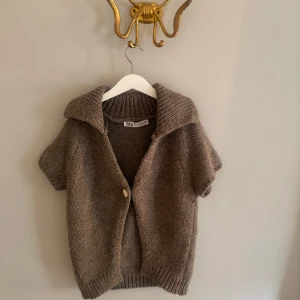 Brun stickad kofta från Zara - Kortärmad brun stickad kofta från Zara med stor krage och en synlig knapp framtill. Koftan har ribbade muddar och en loose passform, perfekt att slänga över en t-shirt eller topp för en chill look.🫶🏼