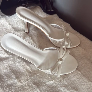 Vita spetsiga pumps med rosett - Eleganta vita pumps med spetsdetaljer och söt rosett framtill. Skorna har öppen tå, smal hög klack och en feminin design. Perfekta för dig som vill ha en chic och stilren look. Materialet är en mix av syntet och spets. Aldrig använda (storlek 38)