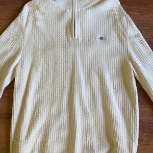 Beige half zip tröja från GANT - Säljer en beige långärmad tröja från GANT i ekologisk bomull. Tröjan har ett snyggt rutigt strukturmönster, ribbad krage och half zip-dragkedja. Liten GANT-logga broderad på bröstet. Perfekt för en clean och stilren look. Endast använd 1 gång
