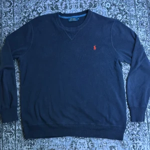 Mörkblå tröja från Polo Ralph Lauren - Snygg mörkblå tröja från Polo Ralph Lauren med klassisk röd broderad logga på bröstet. Tröjan har rund halsringning, ribbade muddar och är långärmad. Perfekt för dig som gillar stilren och tidlös design.