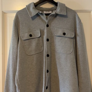 Grå overshirt från Only & Sons  - Snygg grå overshirt från Only & Sons i storlek L. Skjortan har klassisk krage, två stora bröstfickor med lock och svarta knappar hela vägen. Perfekt lager-på-lager-plagg med en clean och stilren look.