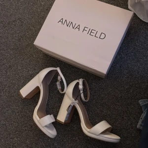 Vita klackar från Anna Field - Stilrena vita klackar från Anna Field med bred rem över tårna och justerbar ankelrem med spänne. Skorna har öppen tå, blockklack och platt sula framtill. Perfekta för dig som vill ha en clean och modern look.