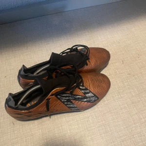 Bronsfärgade fotbollsskor med snörning - Säljer ett par bronsfärgade fotbollsskor från New Balance med svart snörning och svart/vitt mönster på sidan. Skorna har plös i stretchigt material och dobbar för bra grepp på planen. Ovandelen är i syntetmaterial och sulan är svart.