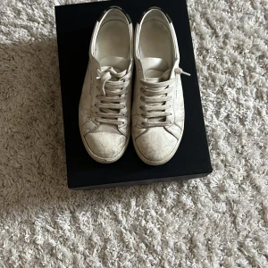 Saint Laurent vita sneakers med camo-detalj - Vita låga sneakers från Saint Laurent med klassisk snörning och rund tå. Skorna har en stilren design i skinn och cool camo-detalj i grönt och brunt på hälen. Perfekta för dig som gillar minimalistisk stil med en twist. Köpta på vestiare för 2300