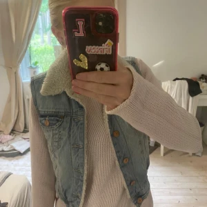 Ljusblå jeansväst med fuskpälsfoder från &Jacket - Säljer en ljusblå jeansväst från &Jacket med mjukt vitt fuskpälsfoder och krage. Västen har bruna knappar framtill och två bröstfickor med lock. Perfekt för lager-på-lager och ger en cool vintagekänsla.
