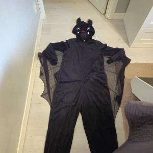 Svart fladdermusdräkt från Divided XS/S - Cool svart fladdermusdräkt från Divided med huva som har stora ögon, öron och tänder. Dräkten har dragkedja framtill och vingar i transparent mesh mellan ärmarna och sidorna. Perfekt för halloween eller maskerad!