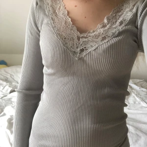 Grå ribbad topp med spets från Style Butler - Supersnygg ljusgrå ribbad topp från Style Butler med v-ringning och spetsdetaljer vid hals och ärmslut. Långärmad och figurnära passform, perfekt att styla med jeans eller kjol. Söt volangkant nertill för extra detalj.
