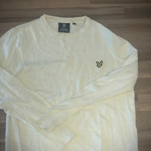 Ljusbeige stickad tröja Lyle & Scott - Snygg ljusbeige stickad tröja från Lyle & Scott med klassisk rund hals och broderad logga på bröstet. Tröjan har långa ärmar och ribbade muddar för en clean look. Perfekt för dig som gillar stilrena och enkla plagg.