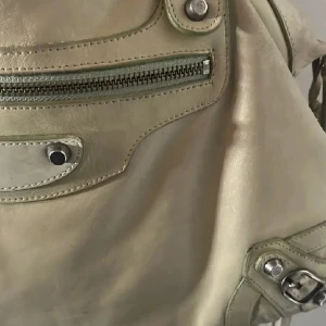 Beige Balenciaga handväska i skinn - Säljer en beige Balenciaga handväska i mjukt skinn med silverfärgade metalldetaljer, dragkedja framtill och dekorativa nitar. Väskan har en klassisk, lite slouchy form och långa tofsar på dragkedjorna. Perfekt för dig som gillar statement-accessoarer. Den är äkta och har serienummer i och stämplar. Säljer för bra pris 