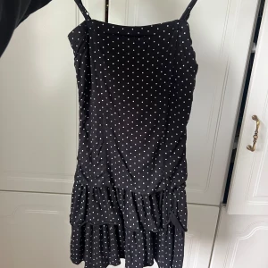 Svart prickig volangklänning - Supersöt svart kortklänning med vita prickar och volangkant nertill. Smala axelband och lätt, flowy passform som ger en chill vibe. Perfekt för varma dagar när du vill känna dig både söt och bekväm.