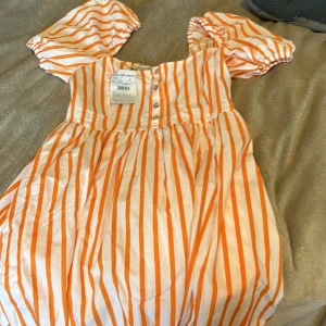 Randig kortklänning från LCW Limited XS - Superfin kortklänning från LCW Limited i vitt och orange med vertikala ränder. Klänningen har puffiga korta ärmar, knappar framtill och en söt, luftig passform. Perfekt för varma dagar och ger en riktigt fräsch sommarkänsla.