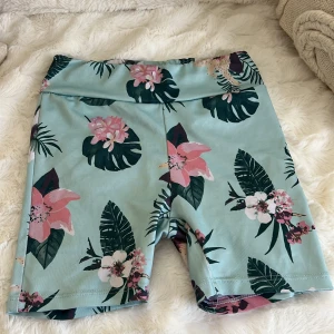 Blommiga träningsshorts från Phelix Sports - Snygga träningsshorts från Phelix Sports i ljusblått med tropiskt blommigt mönster i rosa, vitt och grönt. Modellen är midwaist och har en tight passform, perfekta för träning eller sommarens aktiviteter. Materialet är stretchigt och känns mjukt mot huden.