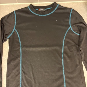 Svart långärmad tröja med blå sömmar - Svart långärmad tröja från 2GUE med kontrasterande blå sömmar. Perfekt för träning eller som baslager under kyliga dagar. Tröjan har en enkel, sportig design och är tillverkad i ett syntetmaterial som andas.