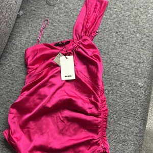 Ceriserosa linnefrån Bik Bok - Säljer en ceriserosa linne från Bik Bok i glansigt satinmaterial. Linnen har en axelbandslös design med draperade detaljer och rynkade sidor. Perfekt för dig som vill sticka ut med färg och snygg passform.