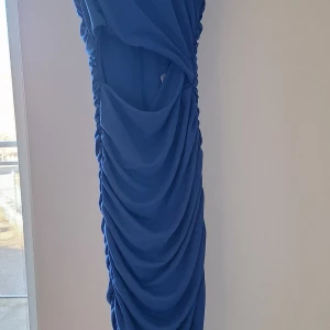 Blå långklänning med drapering från ONLY - Säljer en blå långklänning från ONLY med snygg drapering längs hela klänningen. Klänningen har ärmlös design och en öppen detalj i ryggen. Perfekt för dig som vill sticka ut med en elegant och modern look.