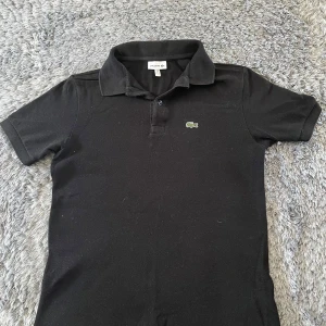 Svart pikétröja från Lacoste  - Klassisk svart pikétröja från Lacoste med korta ärmar och krage. Tröjan har knappar vid halsen och den ikoniska gröna krokodilloggan broderad på bröstet. Perfekt för en stilren och avslappnad look. 