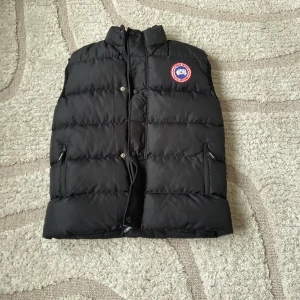 Svart dunväst från Canada Goose - Säljer en svart dunväst från Canada Goose med klassisk logotyp på bröstet. Västen har hög krage, två fickor med dragkedja och tryckknappar framtill. Perfekt för kyliga dagar när du vill hålla värmen utan att kompromissa med stilen.